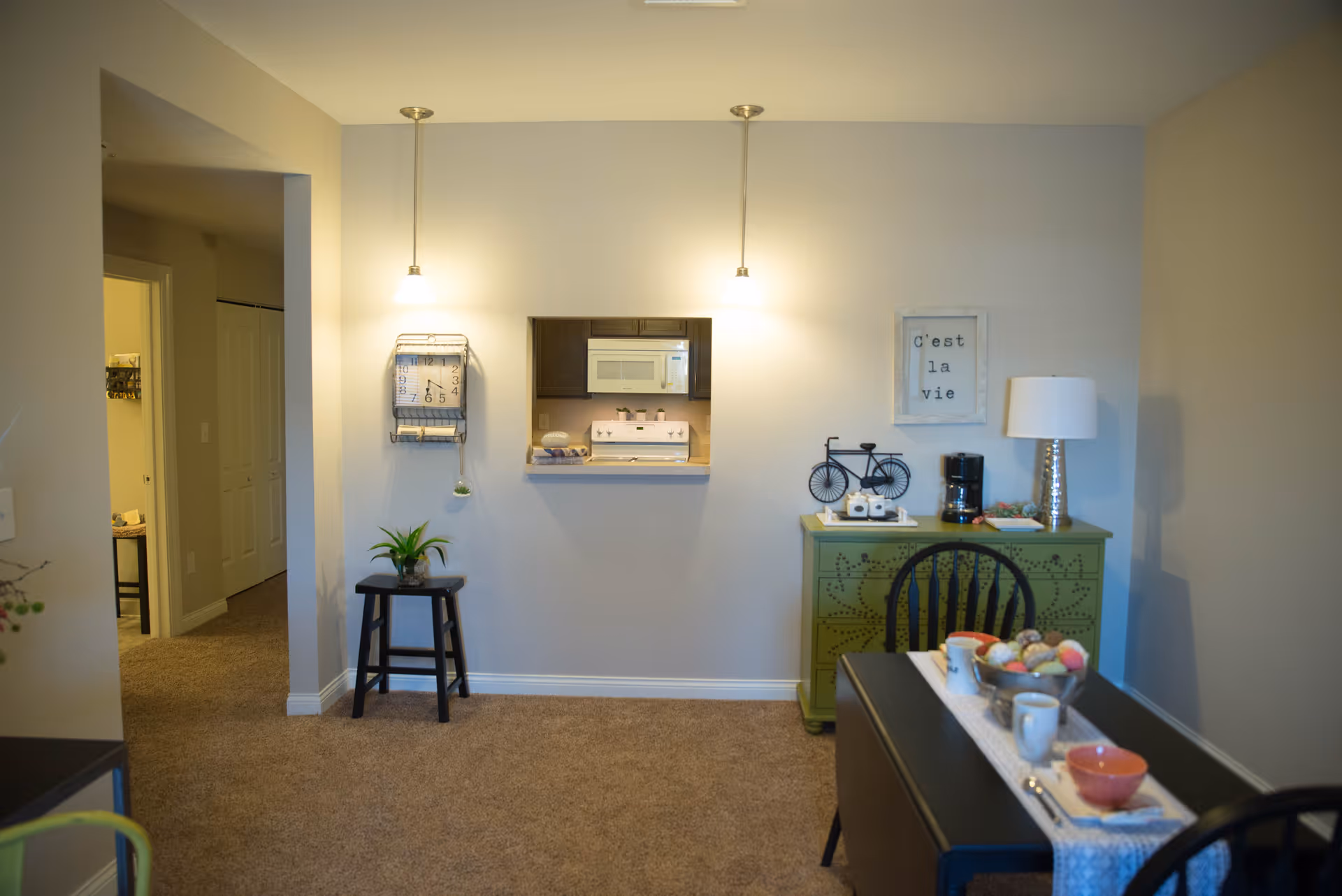 Interior view of a cozy dining area with a small black dining table set for two, a green sideboard with decorative items including a lamp and a coffee maker, and a wall clock and framed art on the wall. The kitchen is visible through a pass-through window above the sideboard.