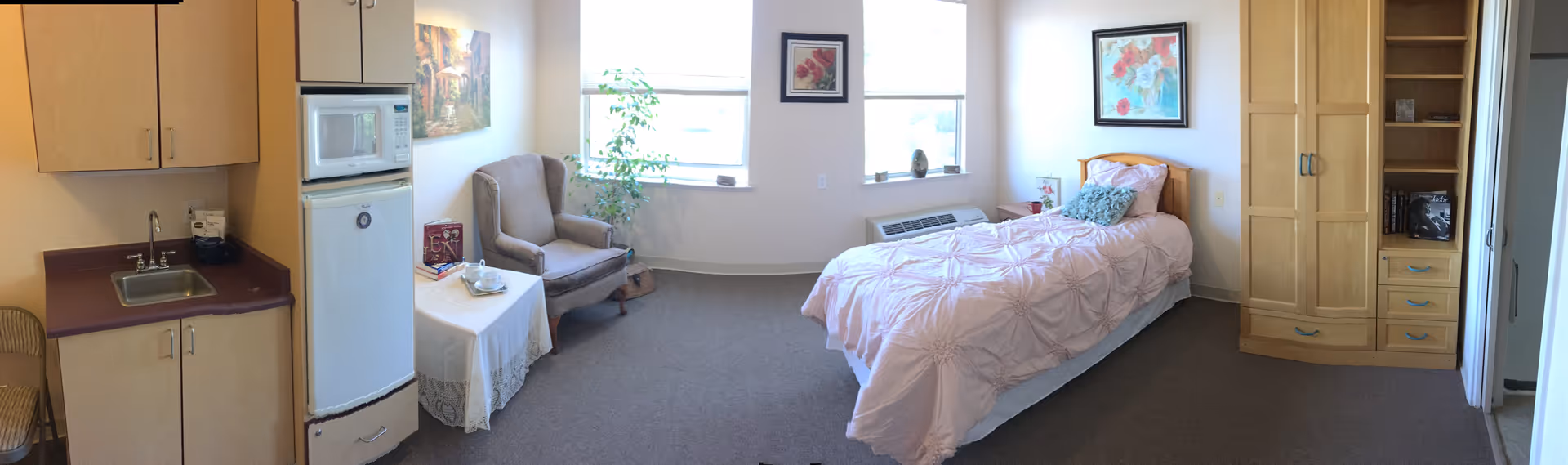 A bright and cozy assisted living room featuring a single bed with a pink comforter, a wooden wardrobe, a bookshelf, and two large windows letting in natural light. There is a comfortable armchair next to a small table with a lace tablecloth, a cup, and some books. A kitchenette area with a sink, microwave, and mini refrigerator is visible on the left side of the room. The walls are decorated with framed floral paintings.