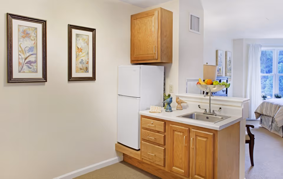 A small kitchenette with wood cabinets, a white refrigerator, a sink and fruit bowl, two framed floral prints on the wall, and a bed visible in the adjoining room.