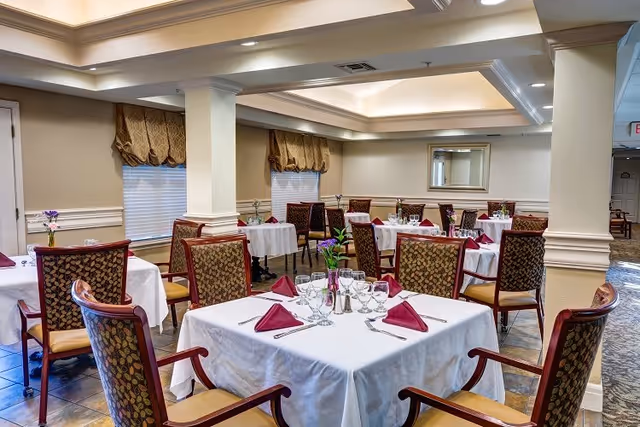 Dining room with multiple tables set with white tablecloths, folded burgundy napkins, glassware, and patterned chairs.