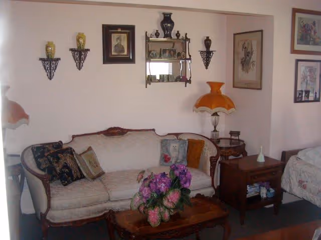 A cozy living room area with a vintage-style beige sofa adorned with decorative pillows. In front of the sofa is a wooden coffee table with a floral arrangement. The walls are decorated with framed pictures and small shelves holding vases and decorative items. To the right, there is a wooden side table with a classic orange lampshade and another small table with books and a small vase. Part of a bed with floral bedding is visible on the right side of the image.