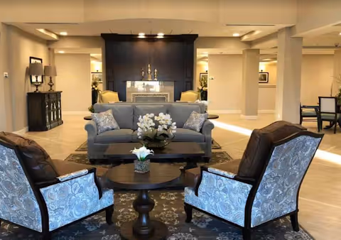 A spacious and well-lit living room area in a senior living facility featuring a gray sofa with patterned cushions, two upholstered armchairs with a floral pattern, a round wooden coffee table with a small flower arrangement, and a fireplace with decorative items on the mantel. The room has beige walls, tiled flooring, and additional seating and furniture in the background.