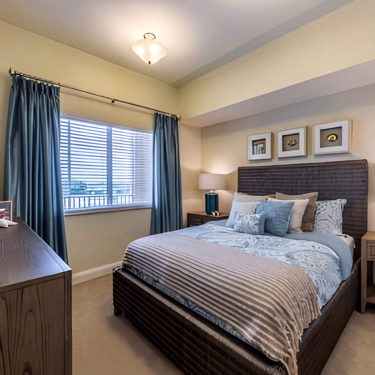 A cozy bedroom with a large bed featuring multiple pillows and a textured blanket. The room has beige walls, a window with blue curtains, a wooden dresser on the left, and a nightstand with a lamp on the right. Three framed pictures hang above the bed's headboard.