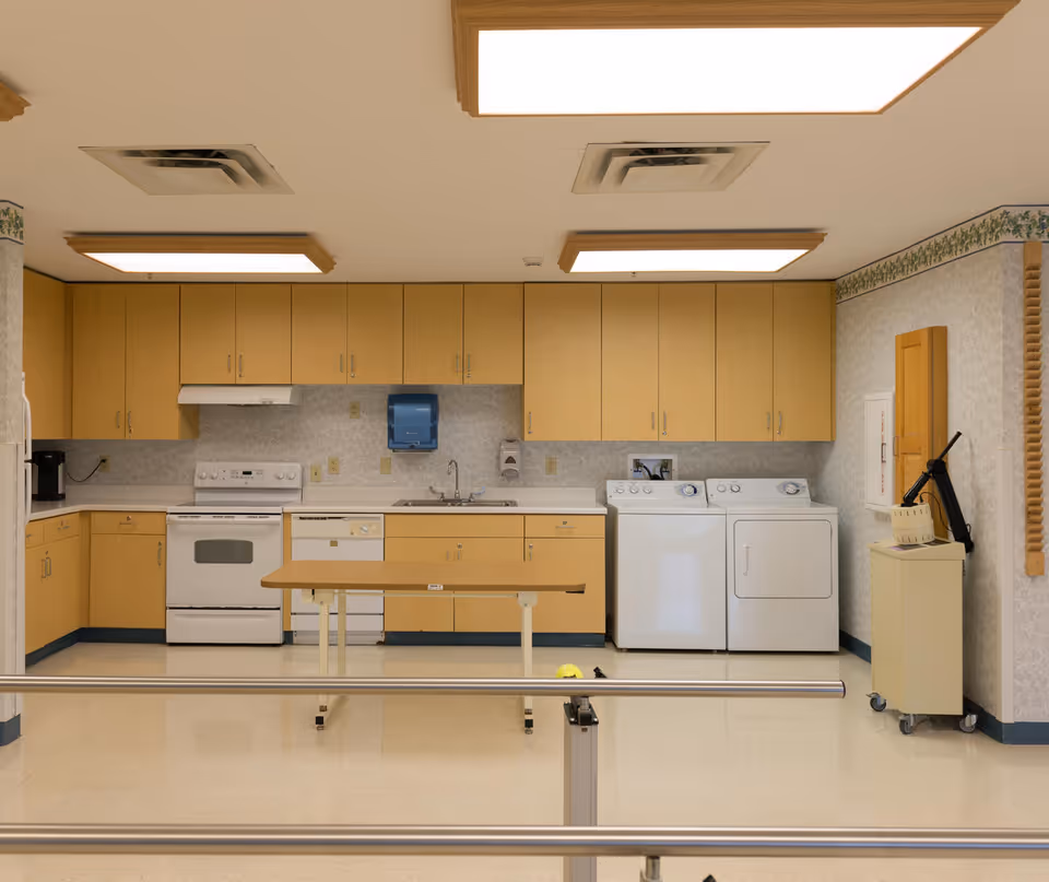 A clean and well-lit room featuring a kitchen area with light wood cabinets, a white stove, dishwasher, and sink. Next to the kitchen appliances are a white washing machine and dryer. The room has a small table on wheels in the center and metal handrails in the foreground. The walls have a light patterned wallpaper with a floral border near the ceiling.