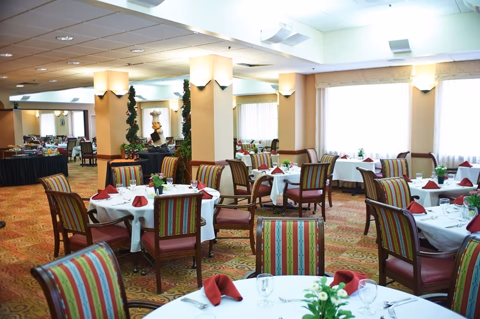 Spacious dining room with round tables covered in white linens, red napkins, striped upholstered chairs, and place settings.