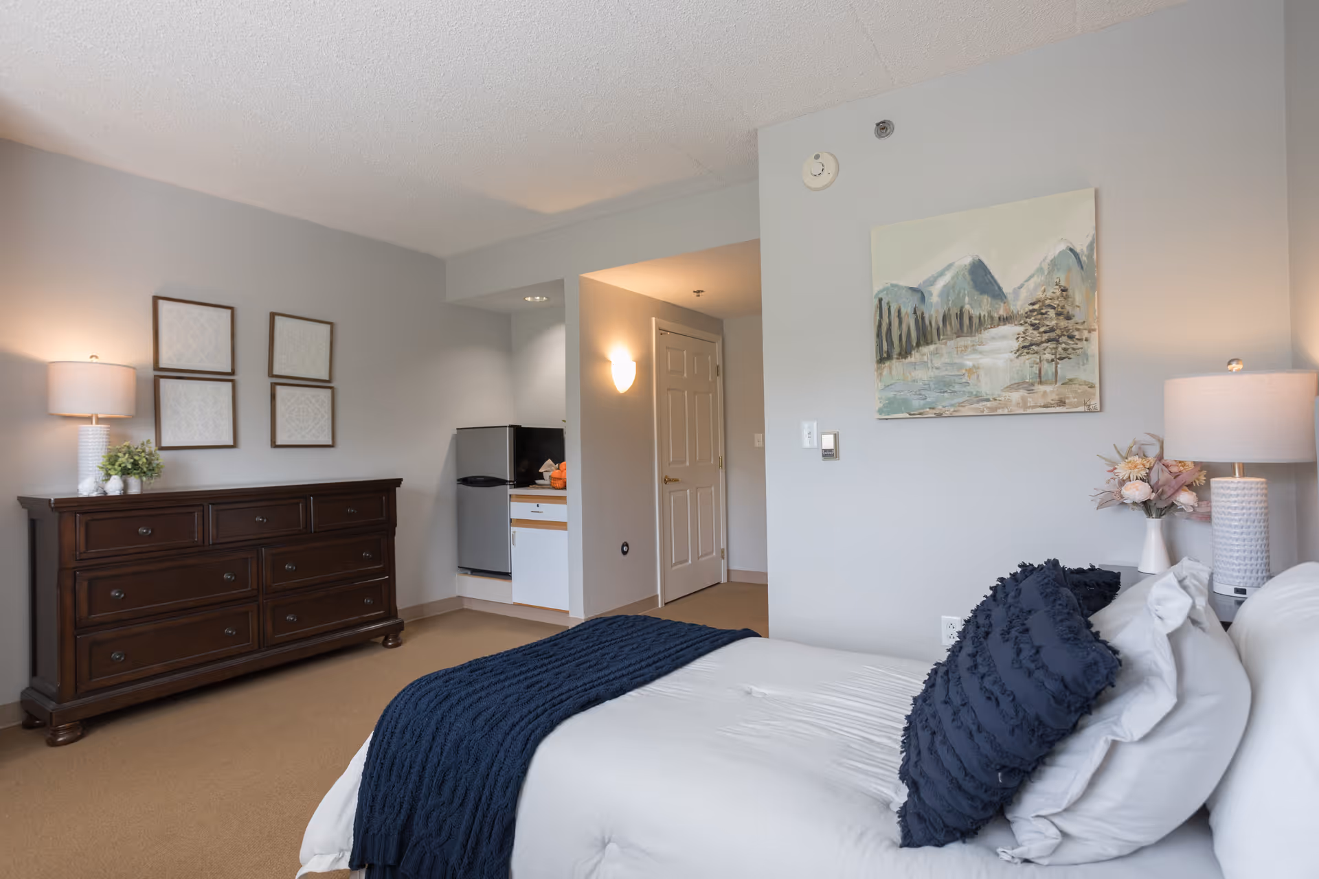 Furnished senior living bedroom with a bed and navy throw, wooden dresser, lamps, wall art and a small kitchenette alcove.