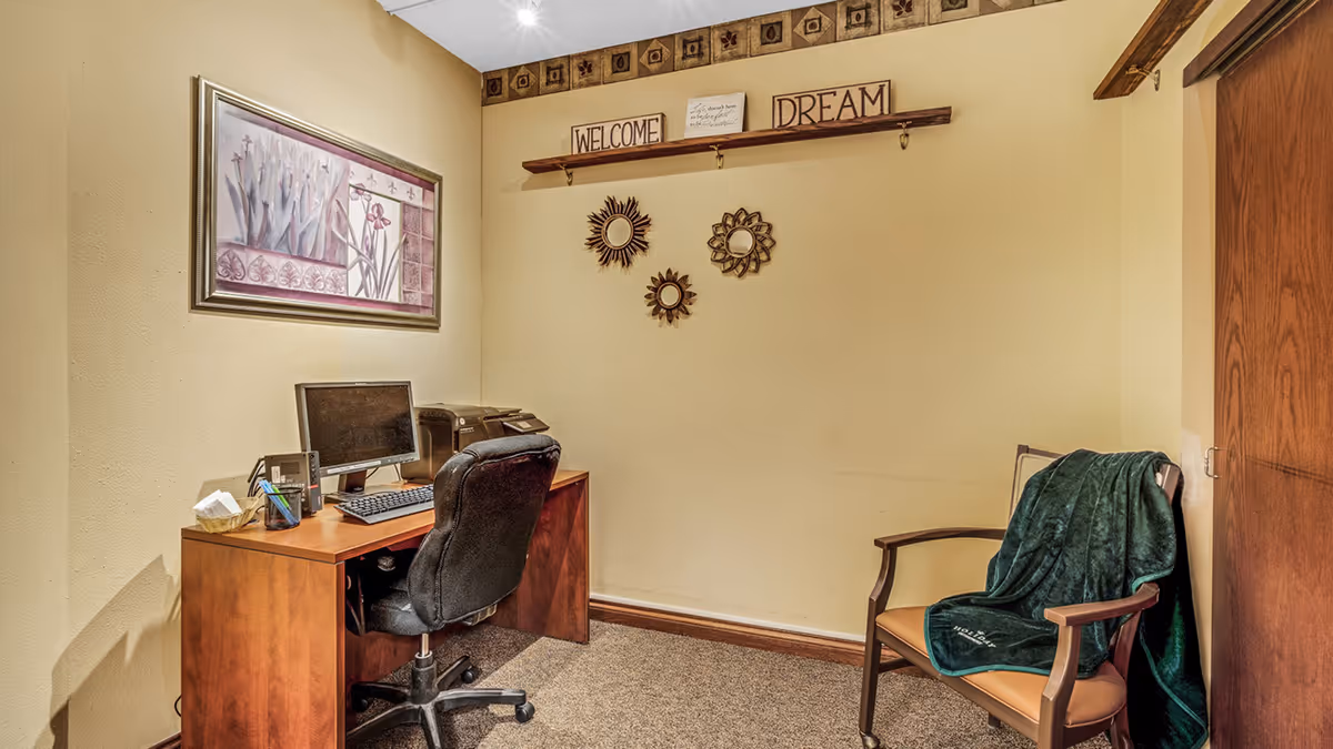 Small interior office nook with a wooden desk, computer and swivel chair, wall decor reading 'Welcome' and 'Dream', and a wooden armchair draped with a green throw.