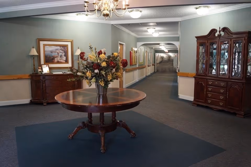 A hallway in a senior living facility with a round wooden table in the center holding a large floral arrangement. The walls are painted light green with a wooden chair rail, and the floor is carpeted. On the left side, there is a wooden sideboard with a framed painting and two lamps. On the right side, there is a large wooden china cabinet with glass doors. The hallway extends into the distance with several doors and framed pictures along the walls, illuminated by ceiling lights and a chandelier above the table.
