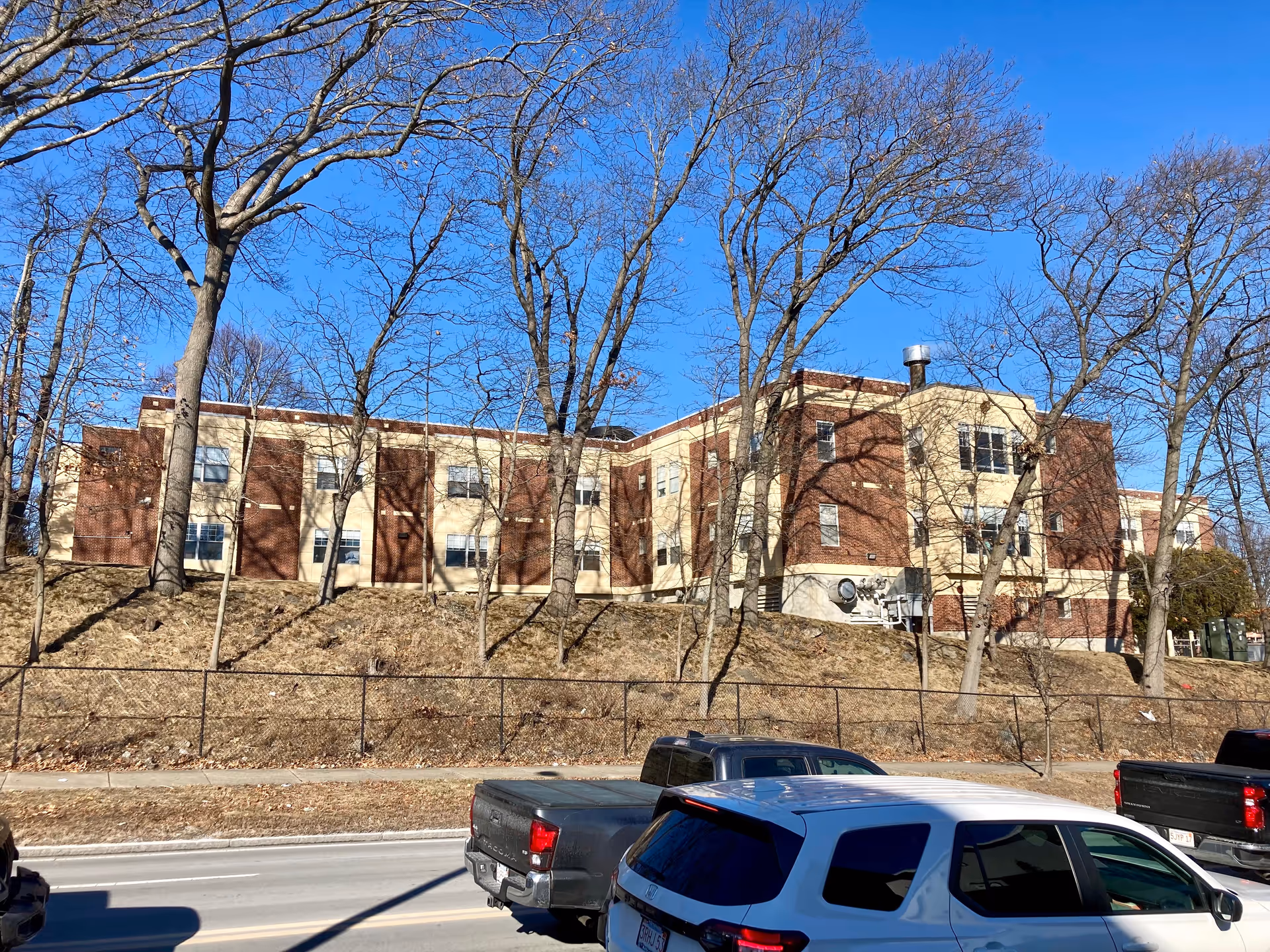 Exterior view of a multi-story brick and beige building behind leafless trees on a clear day with a blue sky. Several parked vehicles are visible along the street in front of the building.