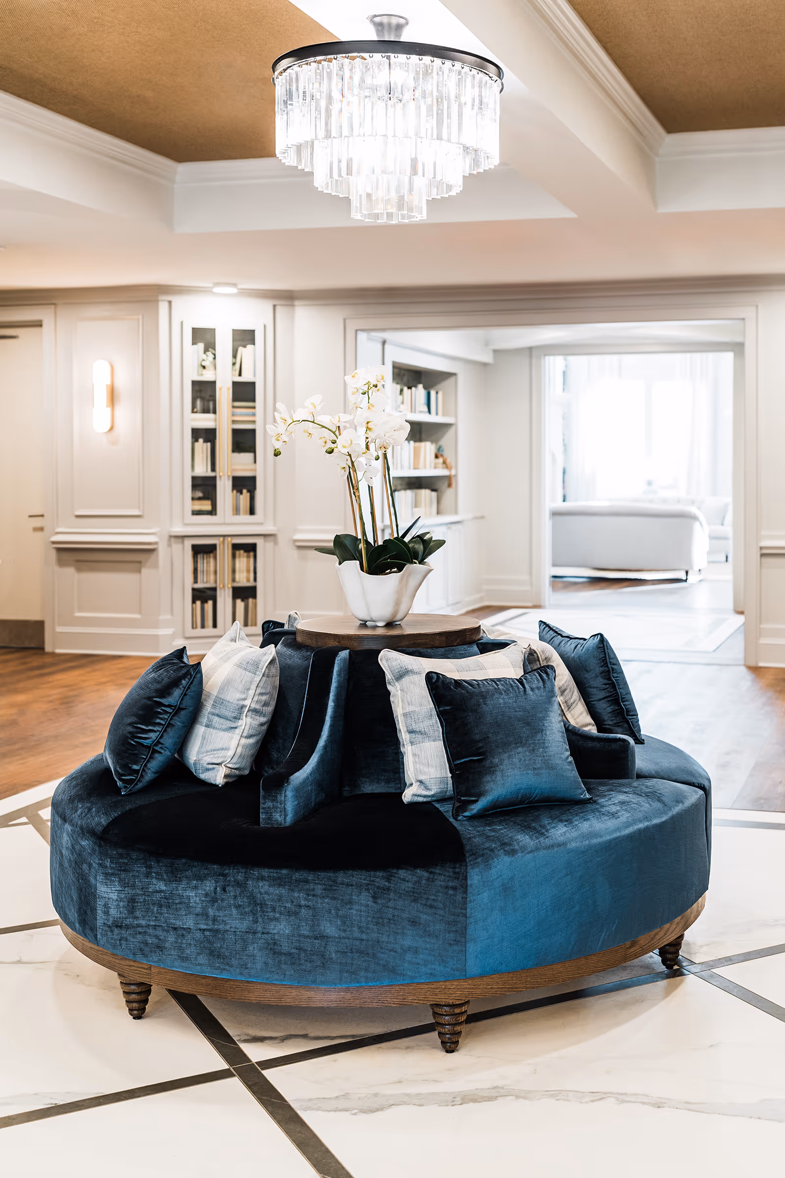 A luxurious circular blue velvet sofa with multiple cushions and a white orchid centerpiece on a wooden table in the middle, set in an elegant room with white walls, built-in bookshelves, wooden flooring, and a crystal chandelier overhead.