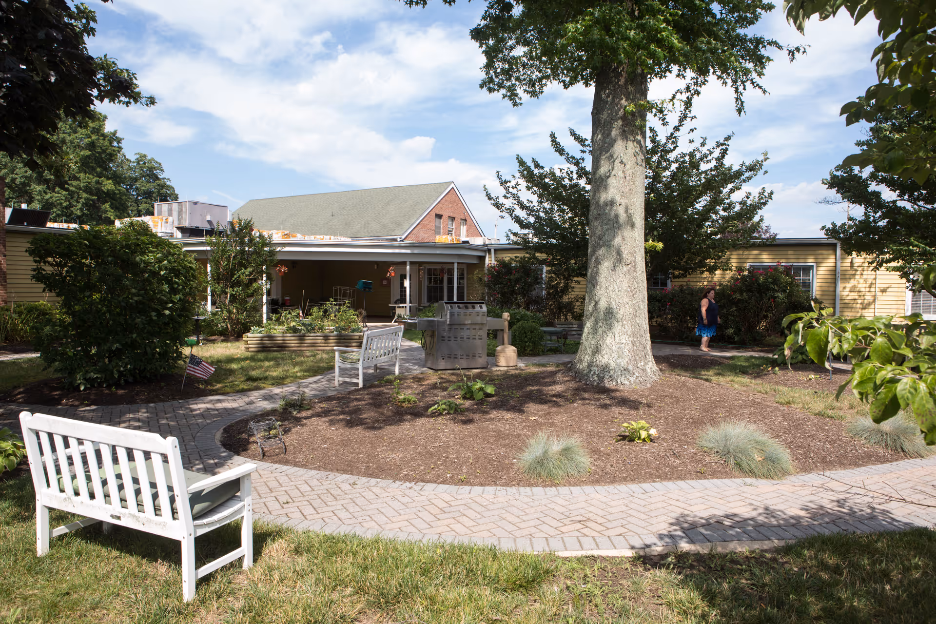 Outdoor garden area at CareOne at Holmdel featuring a large tree in the center surrounded by a circular paved walkway, white benches, green bushes, and a person walking near the yellow building in the background under a partly cloudy sky.