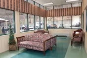 A sitting area inside a senior living facility with large windows along two walls, allowing natural light to fill the room. The room features a floral-patterned sofa, a wooden chair with cushions, a small potted plant, and a green floor. Outside the windows, parked cars and trees are visible.