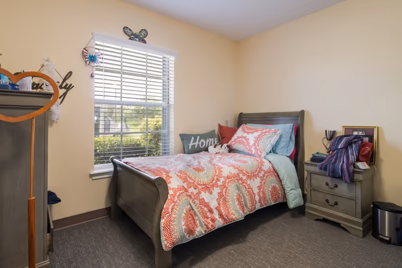 A cozy bedroom with a wooden bed frame and a colorful patterned bedspread in shades of red, orange, and blue. The bed has several pillows, including one with the word 'Home' on it and a small stuffed animal. There is a window with white blinds letting in natural light. Next to the bed is a wooden nightstand with drawers, holding a few items including a framed photo and a folded blanket. The walls are painted a light beige color and the floor is carpeted in a neutral tone.