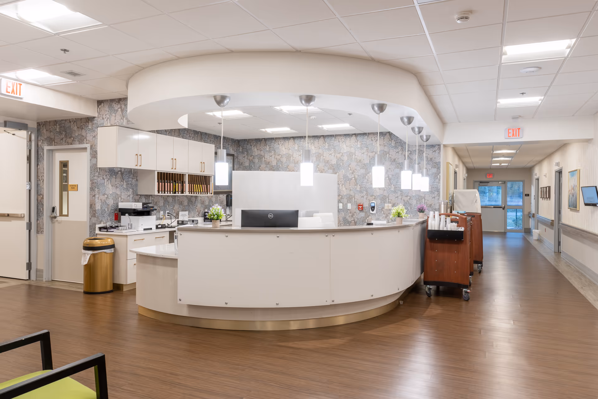 A modern reception desk area in a senior living facility with a curved white counter, hanging pendant lights, and a computer monitor. The background features a patterned wallpaper, cabinets, a printer, and a hallway leading to exit doors. The floor is wooden, and there are small plants on the counter.
