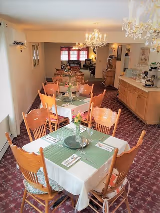 A dining room with several wooden tables and chairs arranged neatly. Each table is covered with a white tablecloth and a green placemat, set with silverware and a small vase with flowers. The room has carpeted flooring, chandeliers hanging from the ceiling, and a sideboard with a coffee maker and other items. In the background, there is a seating area with sofas and windows with red curtains.