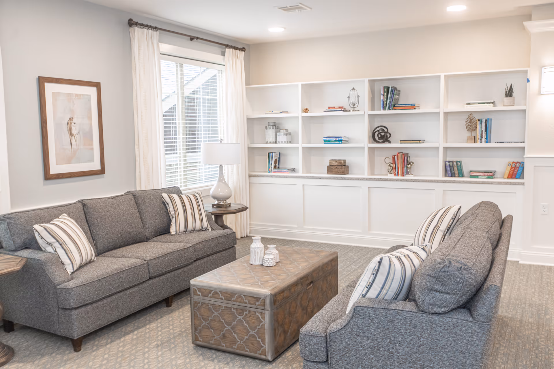 A bright and cozy living room with two gray sofas facing each other, each adorned with striped pillows. Between them is a decorative wooden coffee table with two white ceramic vases. Behind one sofa is a window with white curtains and a table lamp on a side table. The back wall features built-in white shelves with books and decorative items, and a framed picture of a horse hangs on the adjacent wall.