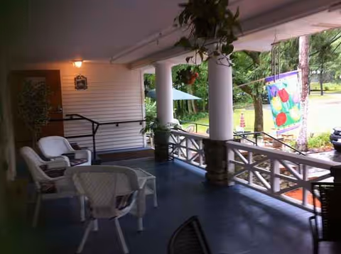 Shaded covered porch with plastic chairs and railing overlooking a grassy outdoor area.
