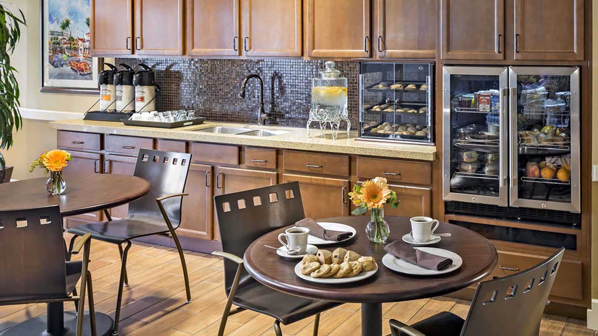 A cozy dining area with round wooden tables and metal chairs. One table is set with plates, cups of coffee, napkins, and a plate of cookies. The background features a countertop with a double sink, coffee dispensers, a glass container of lemon water, a display case with baked goods, and a glass-front refrigerator stocked with various food items. There is a framed picture on the wall and a small vase with a yellow flower on each table.
