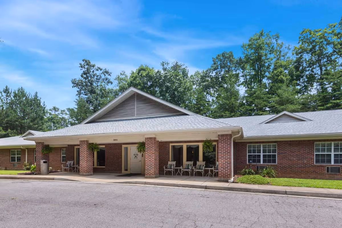 Front exterior view of a single-story brick building with a covered entrance supported by brick columns. There are several chairs arranged under the covered area. The building is surrounded by green grass and trees under a blue sky with some clouds.