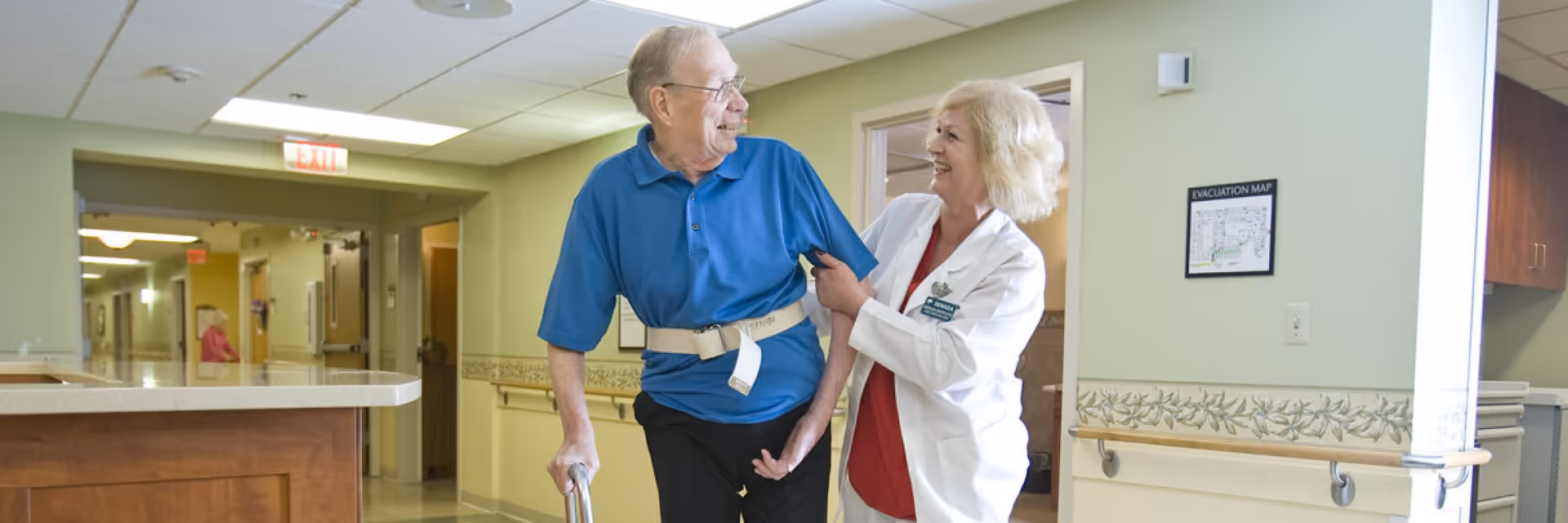 An elderly man using a walker is assisted by a smiling female healthcare worker in a hallway of a senior living facility. The hallway has light green walls, handrails, and an evacuation map on the wall.