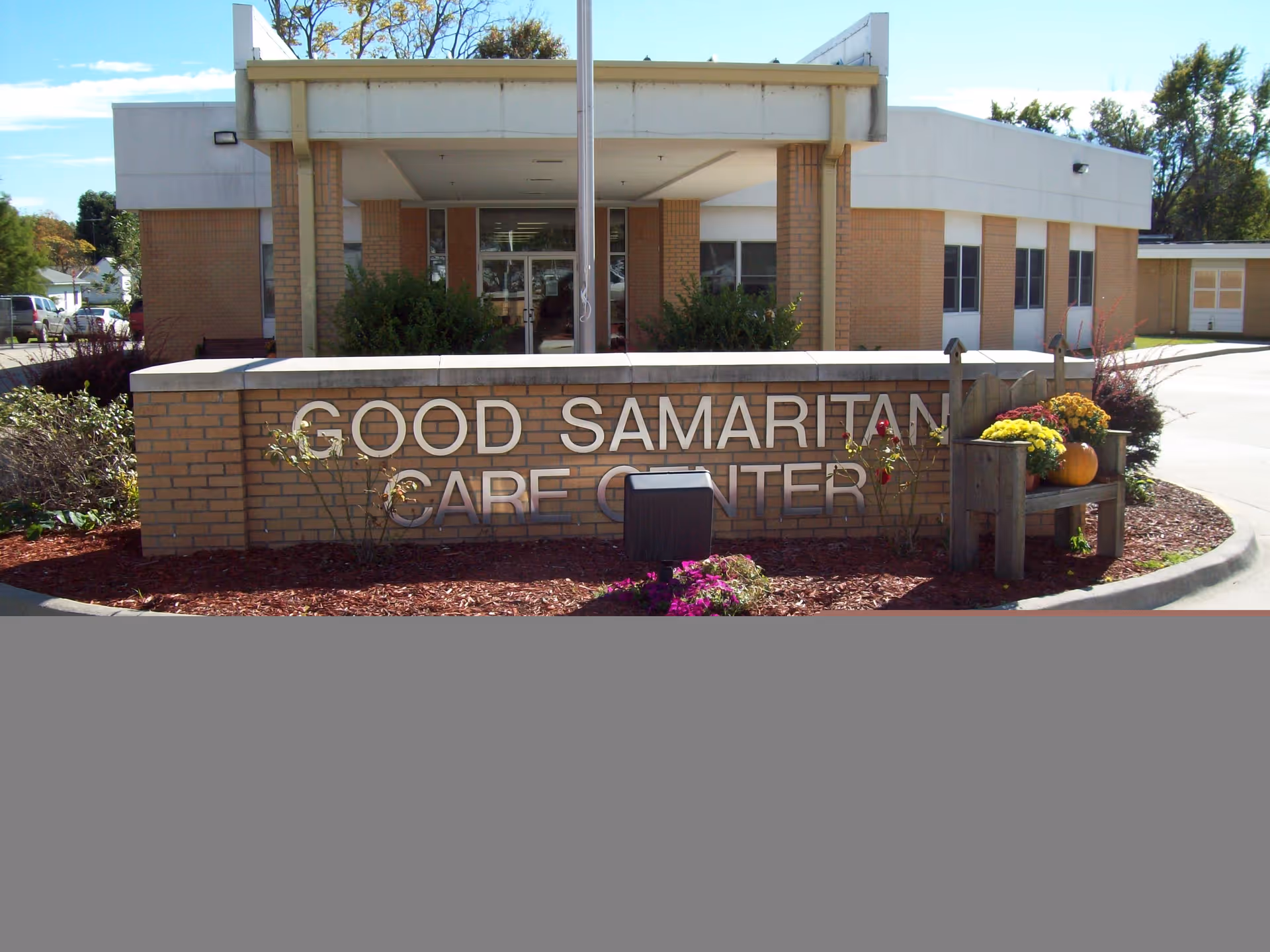 Brick entrance sign reading "Good Samaritan Care Center" in front of a single-story care facility building with landscaping and flowers.