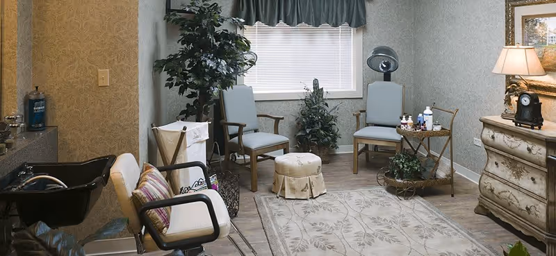 A cozy salon room with two light blue chairs, a small round ottoman, a rolling cart with nail polish and supplies, a vintage-style dresser with a lamp and clock, a black hair washing sink with a chair, and decorative plants. The room has patterned wallpaper and a window with blinds and a valance.
