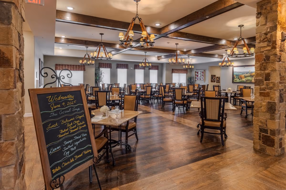 A spacious dining room with multiple tables and chairs arranged neatly. The room features wooden floors, stone pillars, and several chandeliers hanging from a ceiling with wooden beams. A chalkboard menu stands near the entrance displaying the date Wednesday, June 4, 2025, and the meals for the day including Thai Coconut Soup, Balsamic Glazed Pork Chop, Baked Sweet Potato, Roasted Brussel Sprouts, Shrimp Fried Rice, Sautéed Vegetable, Egg Roll, and White Chocolate Bread Pudding. The room is well-lit with natural light coming through several windows with valances.