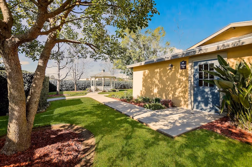 Outdoor garden area at Cottages at Riverside featuring a large tree with mulch around its base, a well-maintained green lawn, a concrete pathway leading to a yellow building with a blue door, and a white gazebo in the background surrounded by trees and hedges.