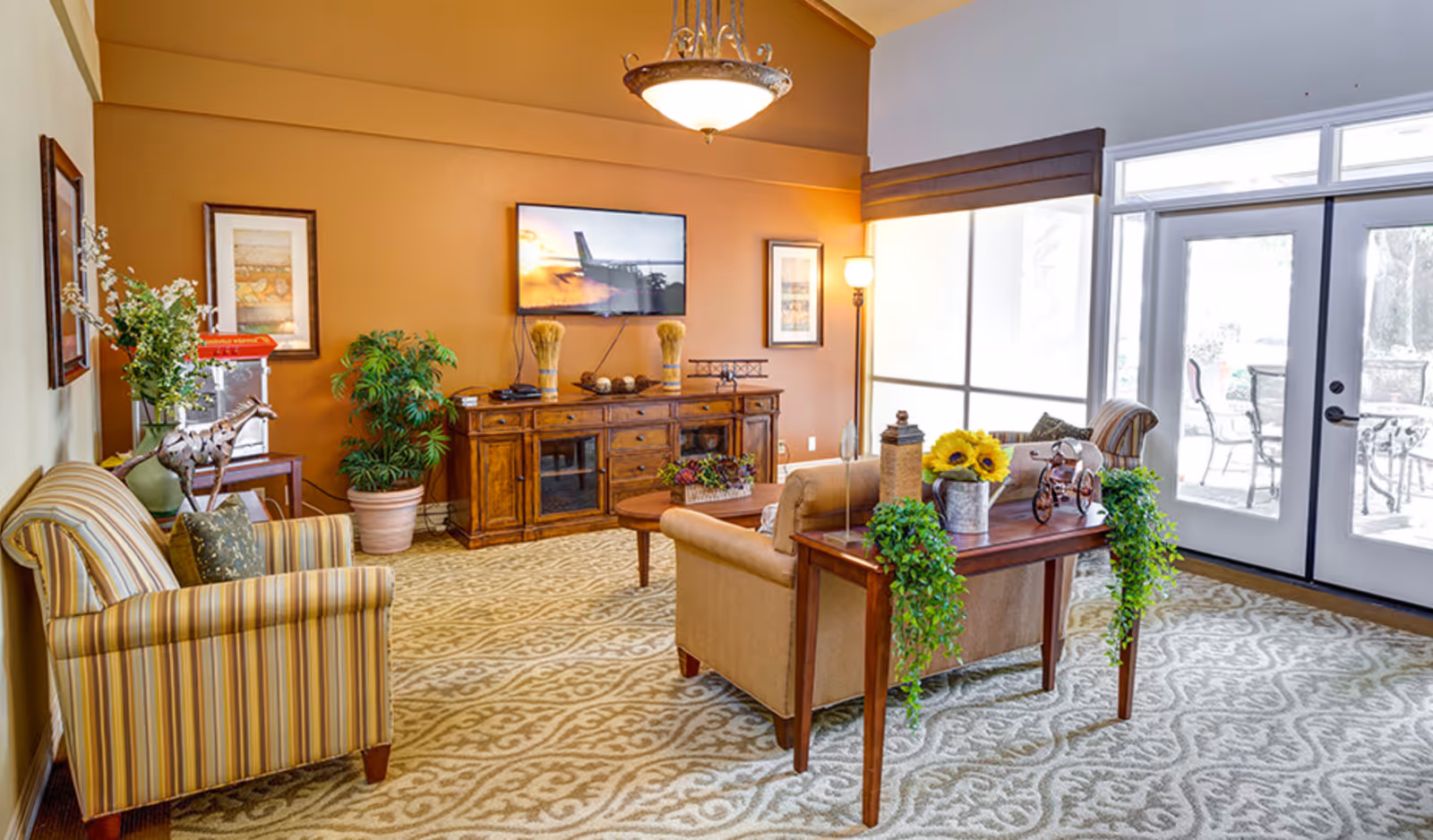 A cozy living room with patterned carpet, a striped armchair, a beige sofa, and a wooden coffee table decorated with plants and sunflowers. A wooden entertainment console holds decorative items and a flat-screen TV mounted on a warm orange wall. Large windows and glass doors let in natural light and lead to an outdoor patio with chairs and a table.