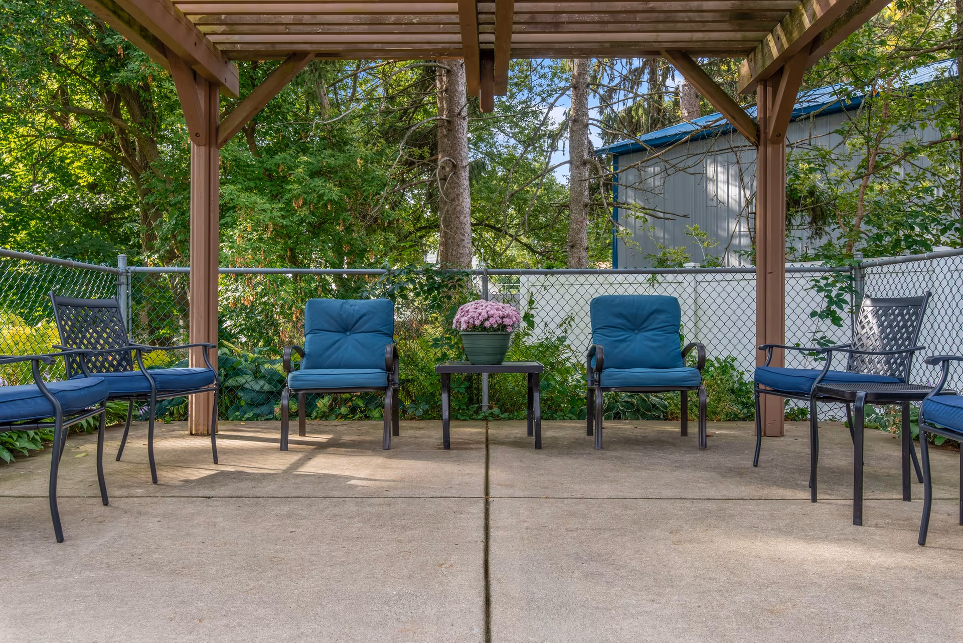 Outdoor covered patio area with six chairs arranged around a small table holding a potted plant with pink flowers. The patio is surrounded by a chain-link fence and greenery, with trees and a building visible in the background.