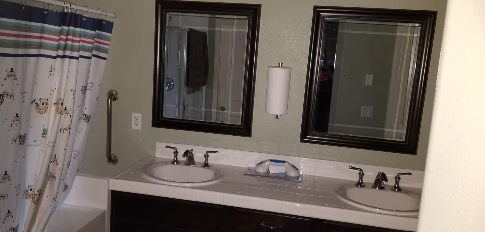 Bathroom with two sinks, each with a mirror above it. There is a paper towel holder mounted on the wall between the mirrors. To the left, a shower curtain with colorful stripes and llama designs is partially visible, along with a grab bar on the wall next to the bathtub.