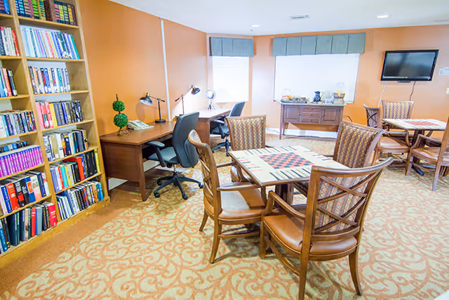 A well-lit room with a patterned carpet featuring two wooden tables with chairs around them. One table has a checkerboard game set up. Along one wall, there are two desks with office chairs, desk lamps, and decorative plants. A bookshelf filled with books is on the left side of the image. The walls are painted a warm orange color, and two windows with gray valances let in natural light. A flat-screen TV is mounted on the wall above a sideboard with decorative items.