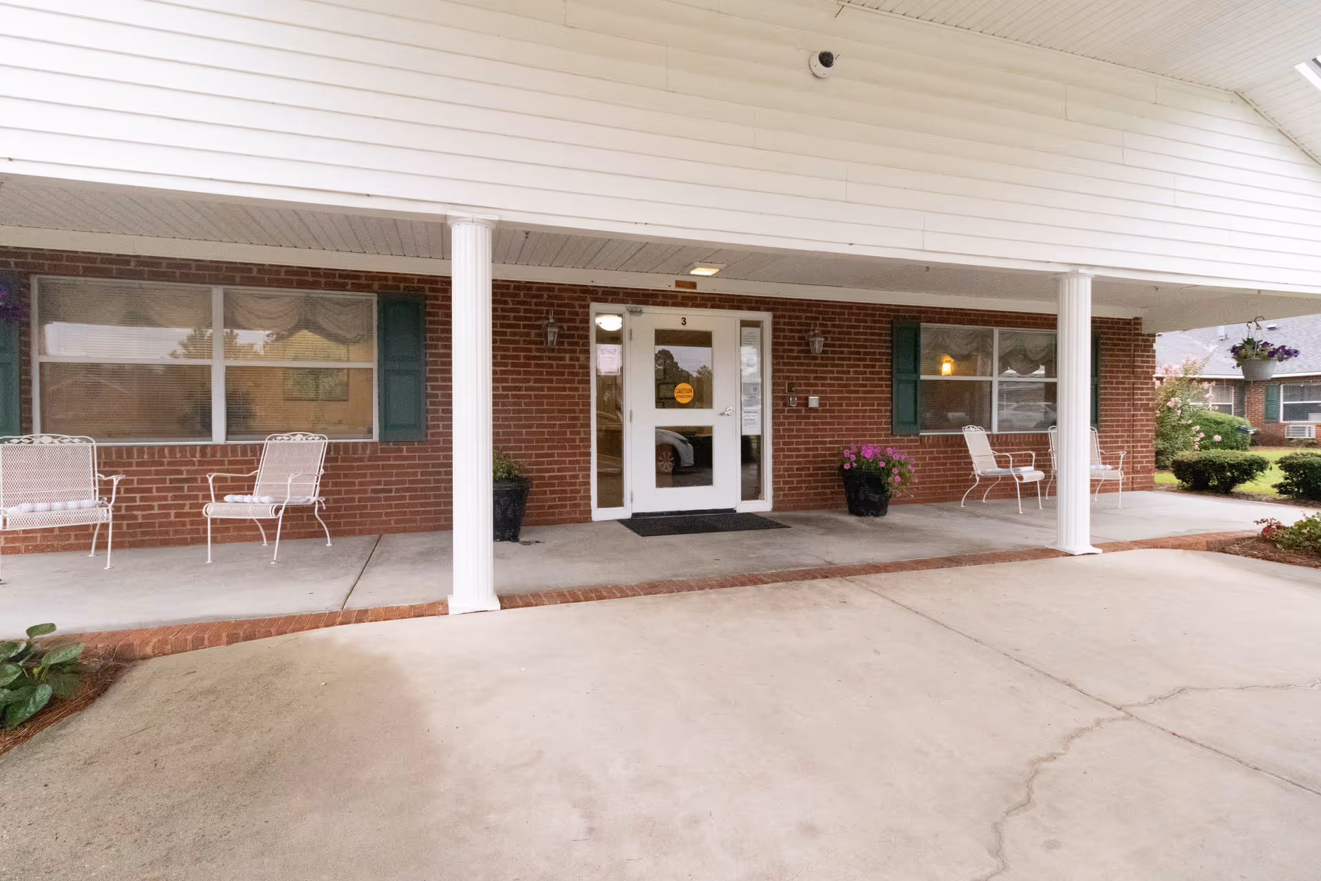 Covered brick front entrance with white columns, a glass door, potted flowers and outdoor chairs on the porch.