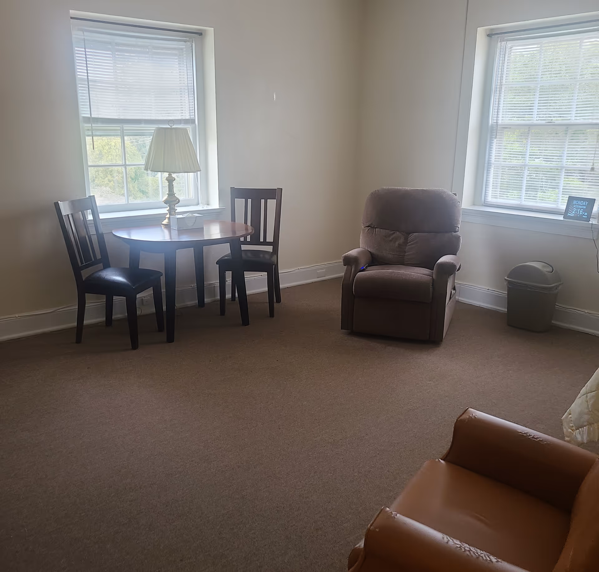 A simple living room with two windows, a round table and two chairs, a lamp, a recliner and an armchair.