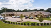 Exterior view of a single-story senior living facility building with a parking lot in front containing several cars. The building has a beige exterior with multiple entrances and a well-maintained lawn with small trees and shrubs.