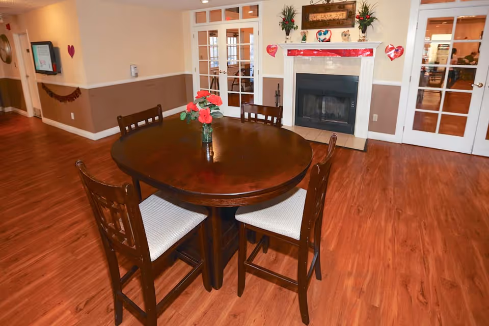 A cozy senior living common area with a round wooden table and four chairs with white cushions. The room features wood flooring, a fireplace decorated with red and pink heart-shaped decorations, and a sign above the fireplace that reads 'Live Laugh Love'. There are double glass doors behind the fireplace and a wall-mounted TV on the left side.