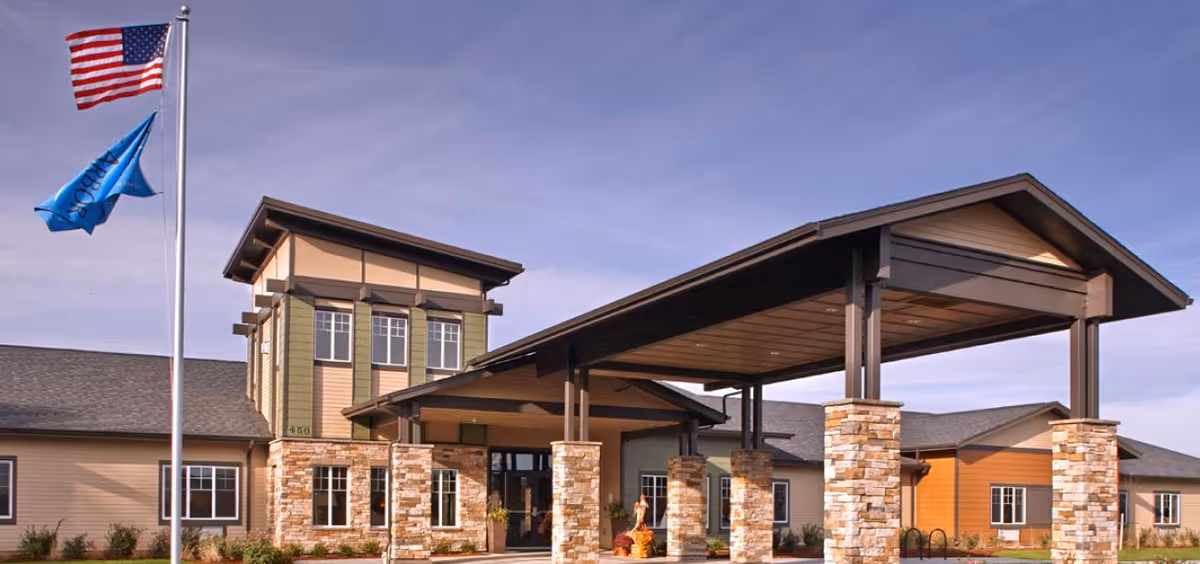 Front entrance of a senior living building with a large covered porte-cochère supported by stone pillars and flagpoles flying an American flag.