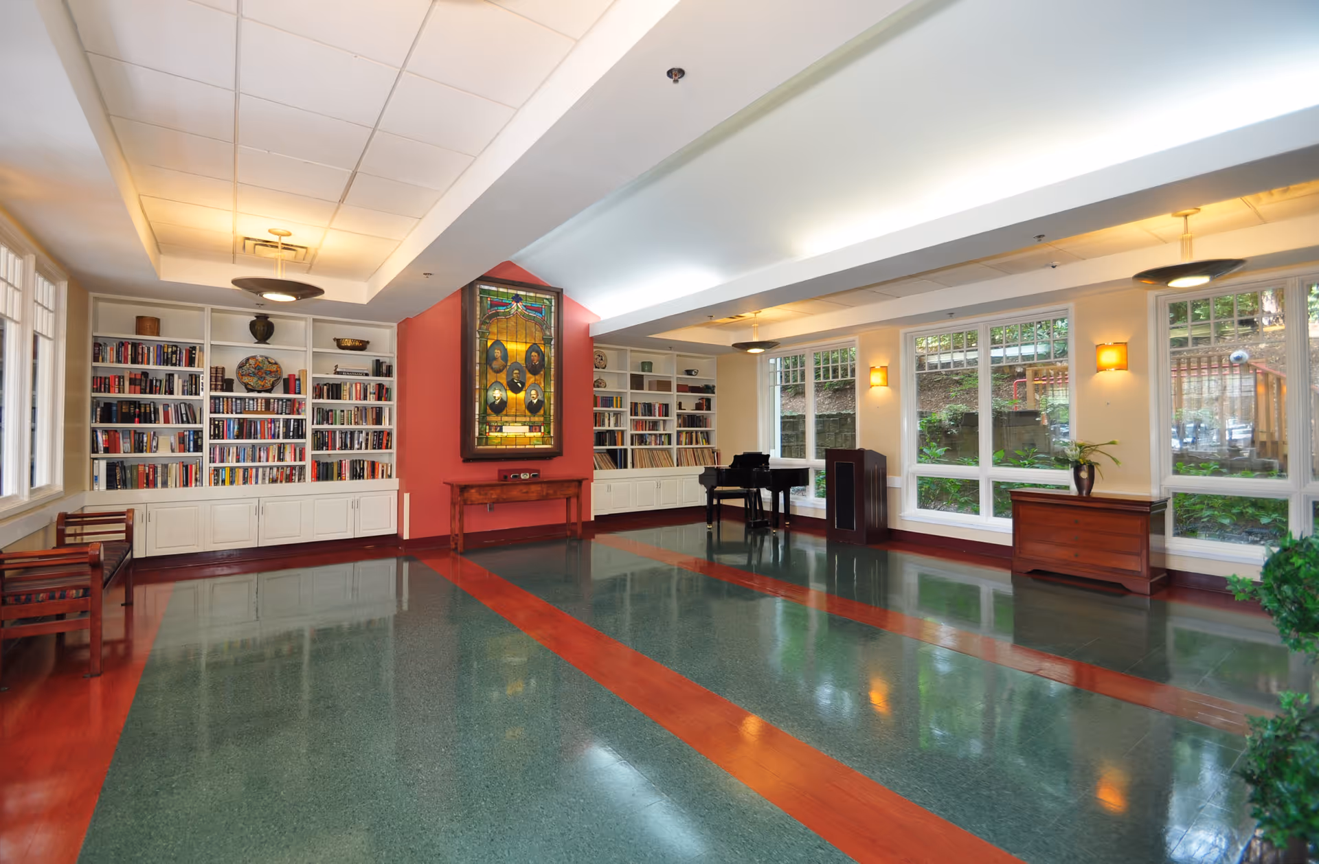 A spacious, well-lit room with large windows showing greenery outside. The room features built-in white bookshelves filled with books, a stained glass artwork on a red accent wall, a black grand piano, wooden benches, and wooden furniture pieces. The floor has a polished green and reddish-brown pattern, and the ceiling has recessed lighting and modern light fixtures.