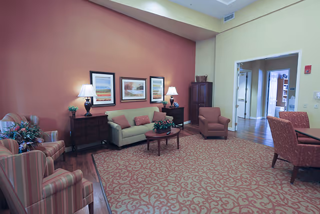 A cozy living room with a patterned carpet, a beige sofa with cushions, two striped armchairs, a wooden coffee table with a plant, two side tables with lamps, framed artwork on a reddish-brown accent wall, and an open doorway leading to another room.