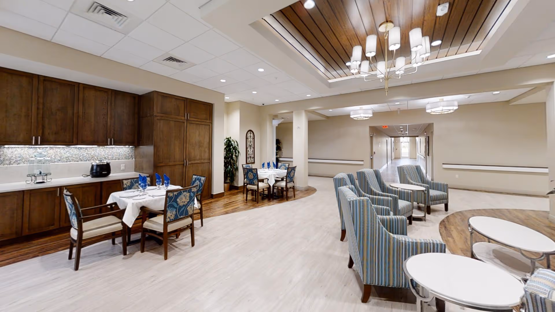 Interior view of a senior living facility common area with a dining section featuring tables set with white tablecloths and blue napkins, wooden cabinets along the wall, and a seating area with striped armchairs and round white tables. The ceiling has recessed lighting and a wooden panel section with a chandelier.