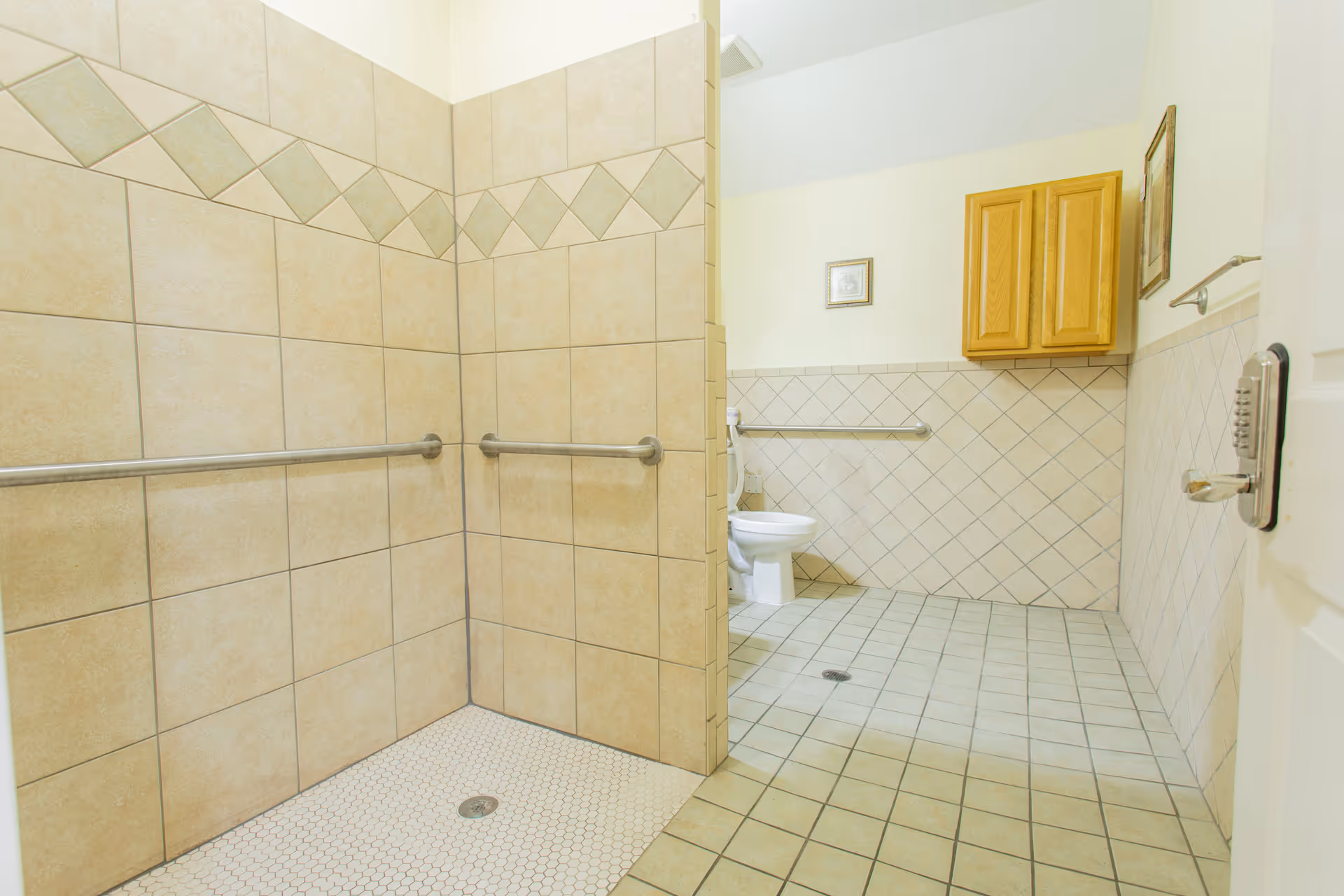 A spacious bathroom with beige tiled walls and floors. The room features a walk-in shower with grab bars on the walls and a drain on the floor. In the background, there is a toilet with a grab bar beside it, a wooden cabinet mounted on the wall, and framed pictures hanging on the walls.