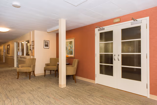 A hallway in a rehabilitation and nursing center with beige walls and carpeted floor. There are two beige armchairs and a small table with a flower vase against a red accent wall. Double white doors with glass panels are on the right side, and framed pictures hang on the walls.