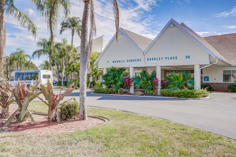 Front entrance of Merrill Gardens at Barkley Place with landscaped palms, a covered entry, and a parked shuttle bus.