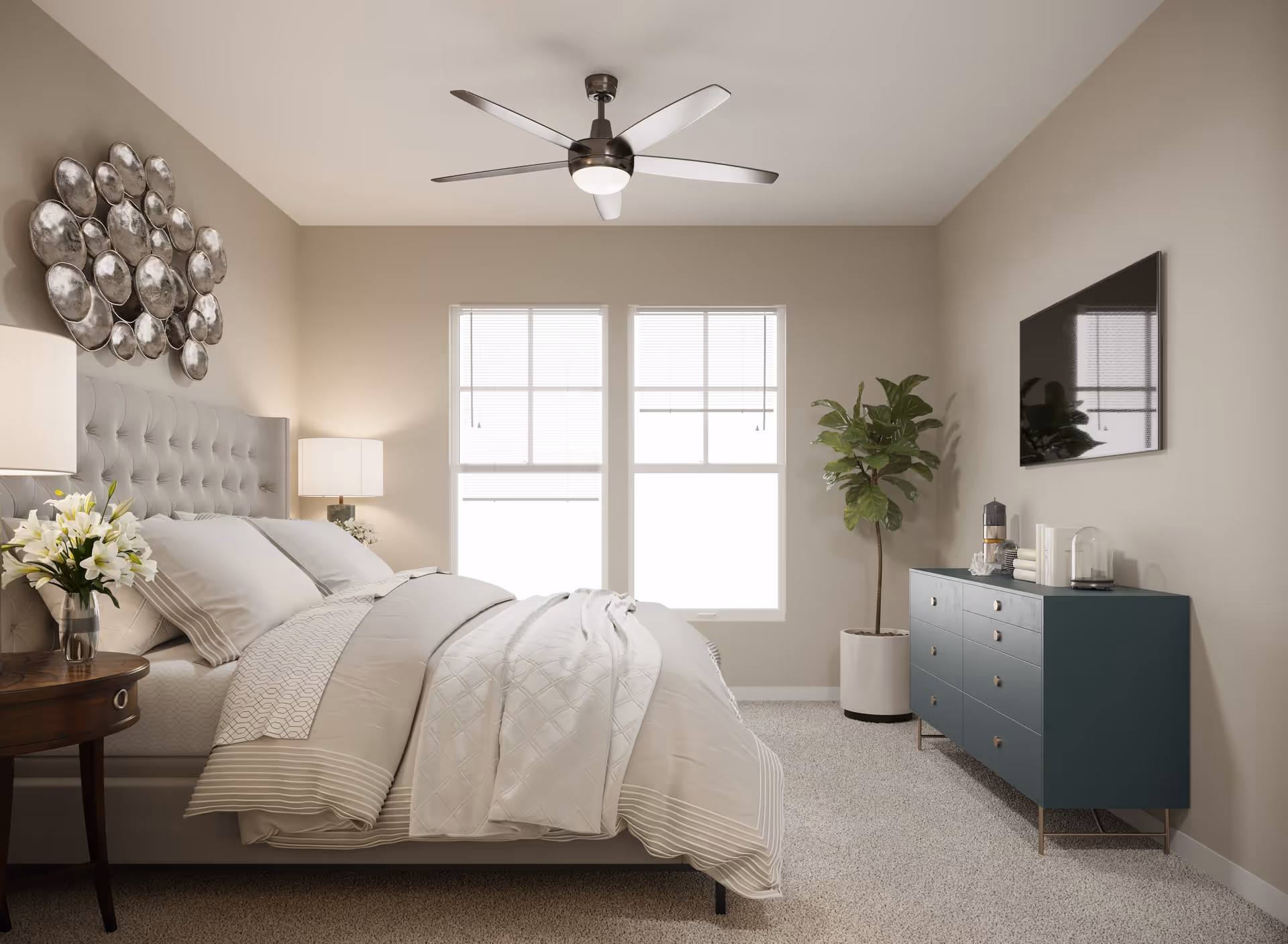 A neatly made bed with white and beige bedding in a bedroom featuring a tufted headboard, two windows with blinds, a ceiling fan with light, a dark teal dresser with decorative items, a potted plant, and a round wooden side table with a vase of white flowers.