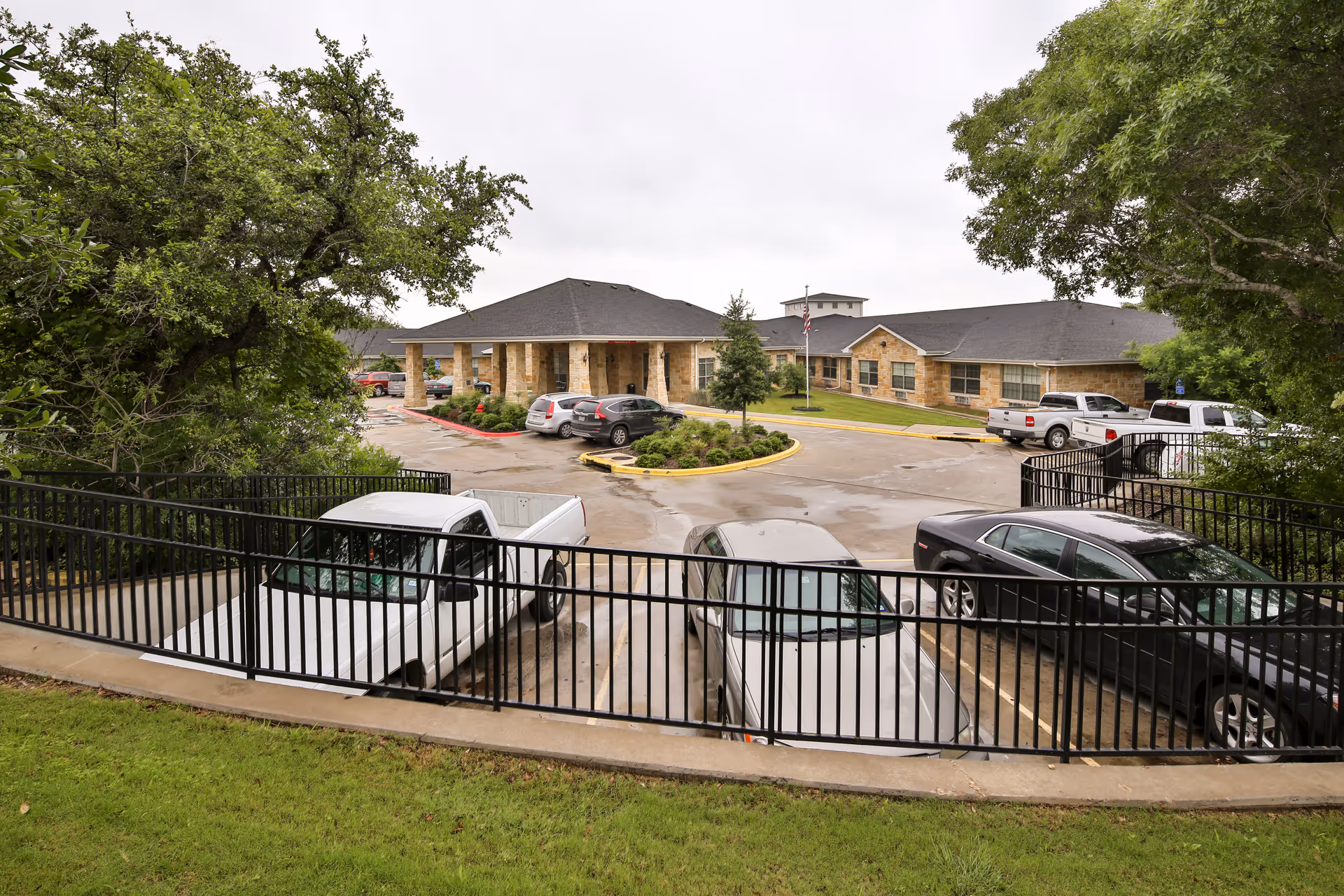 Front exterior of a single-story stone nursing facility with a covered entrance, circular driveway and parked cars seen from behind a black metal fence.