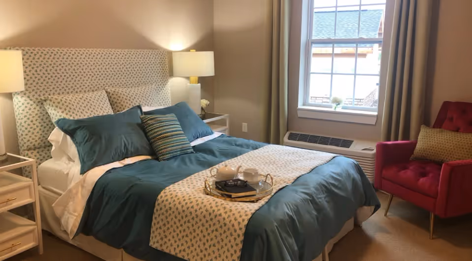 Bright, styled bedroom with a teal-covered bed, patterned headboard, bedside lamps, a tray on the bed, a window, and a red armchair.