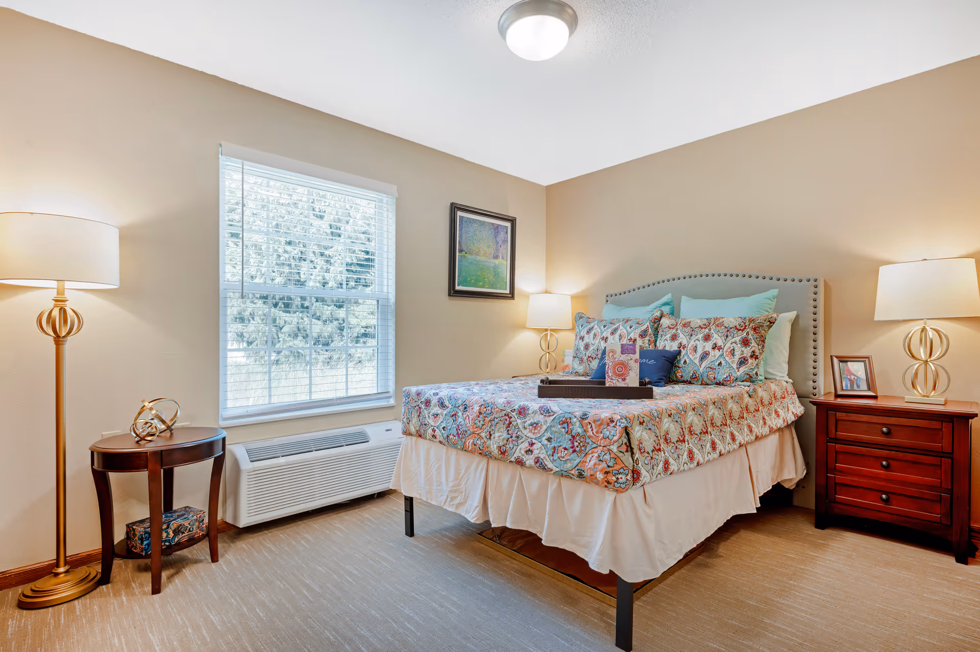 A cozy bedroom with a bed featuring a colorful floral quilt and multiple pillows. There are two bedside tables with lamps on each side of the bed. A window with white blinds lets in natural light, and a framed painting hangs on the wall above the bed. The room has beige walls and carpeted flooring.