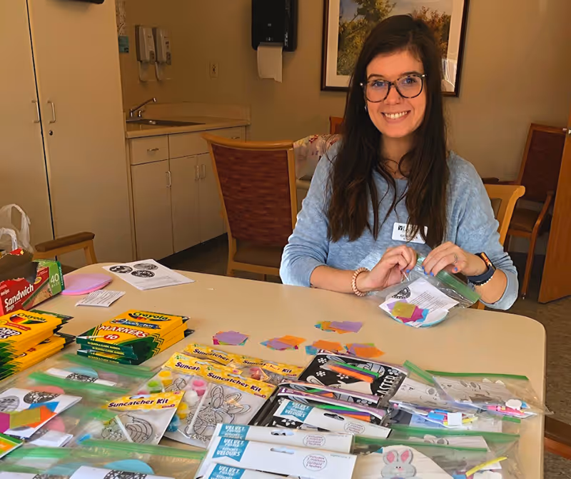 A woman with long brown hair and glasses sits at a table covered with various craft supplies including Crayola markers, Suncatcher kits, and colorful paper pieces. She is smiling and holding a small plastic bag with craft materials. The room has beige walls, a countertop with a sink, and wooden chairs.