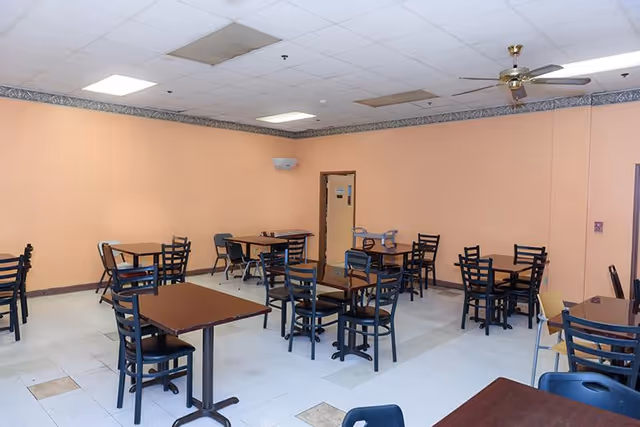 Empty dining room with multiple small tables and chairs on a tiled floor beneath fluorescent lights and a ceiling fan.