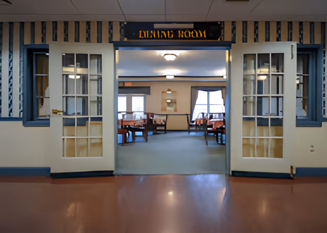 Entrance to a dining room in a senior living facility with double glass-paneled doors open, showing tables and chairs inside. The sign above the entrance reads 'DINING ROOM'.